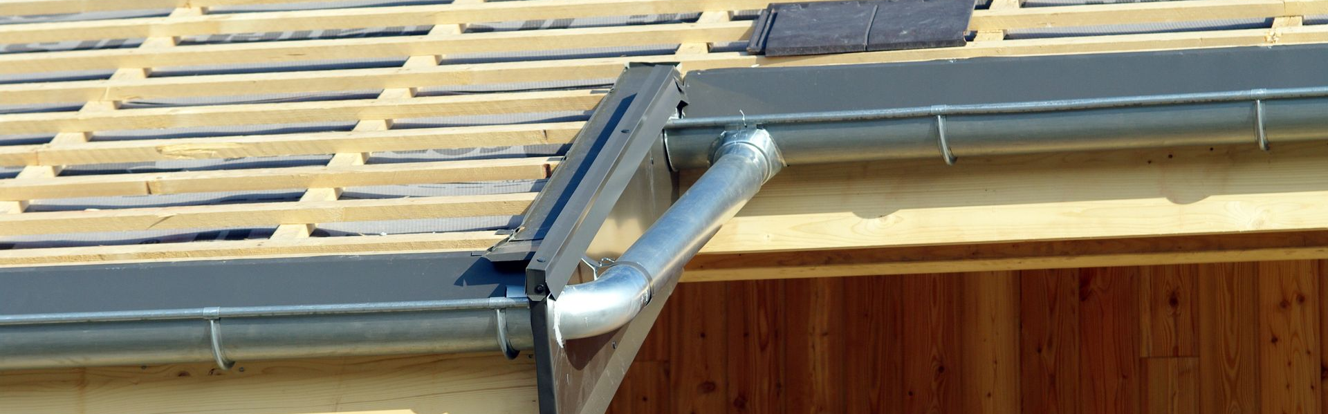 Une gouttière métallique avec une descente pluviale fixée à une charpente de toit en bois.