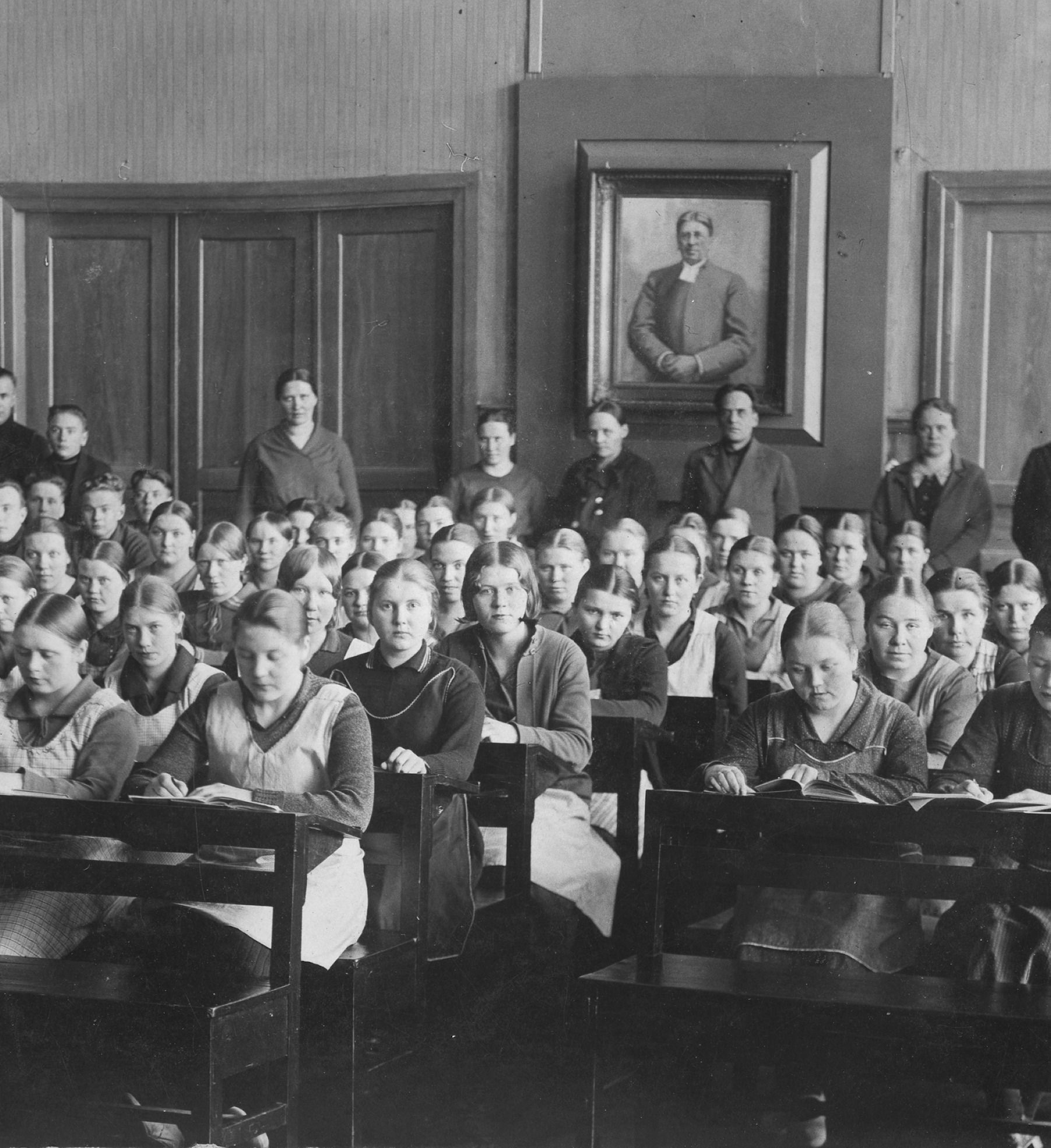 Vintage-mustavalkoinen luokkahuonekuva, jossa oppilaita istuu riveissä pulpettien ääressä kehystetyn muotokuvan alla.