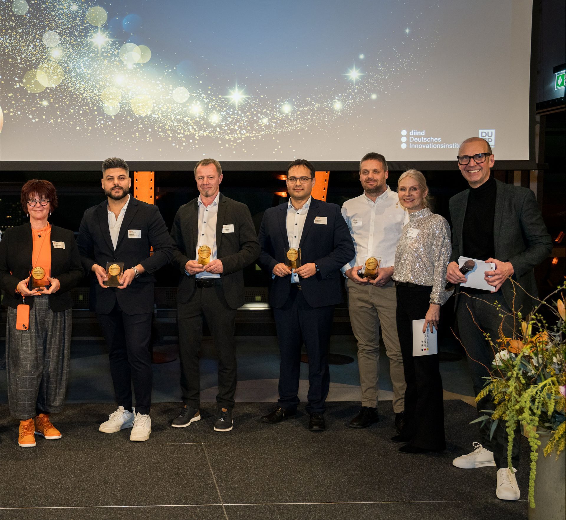 Eine Gruppe von Menschen hält Auszeichnungen auf einer Bühne, vor festlichem Hintergrund mit Lichtern.