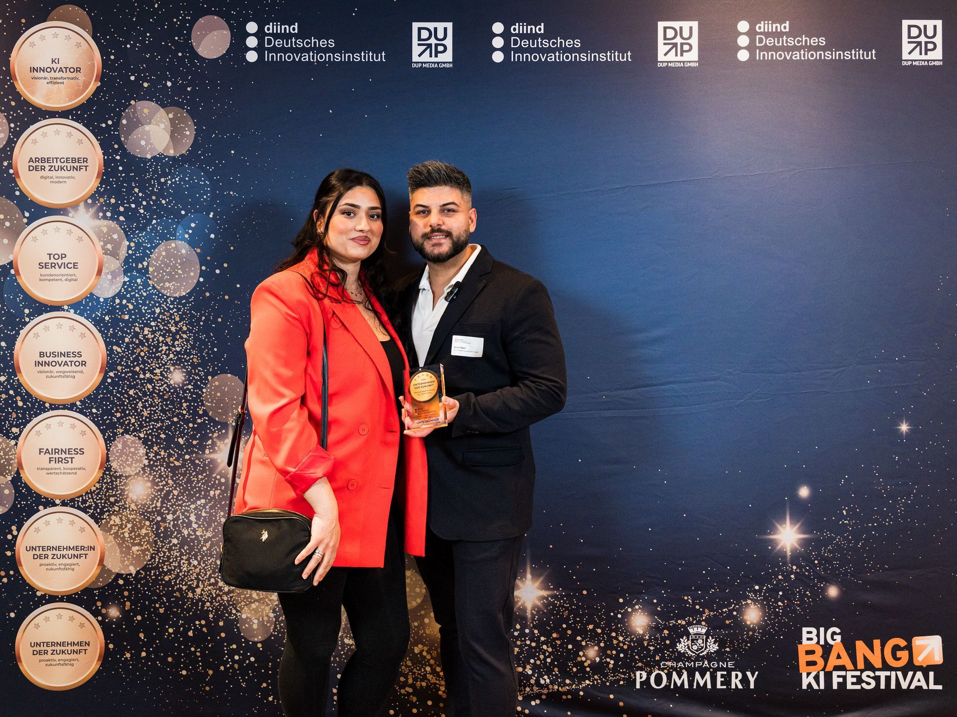 Zwei Personen posieren mit einer Auszeichnung vor blauem Hintergrund. Die Frau trägt einen roten Blazer, der Mann einen Anzug.