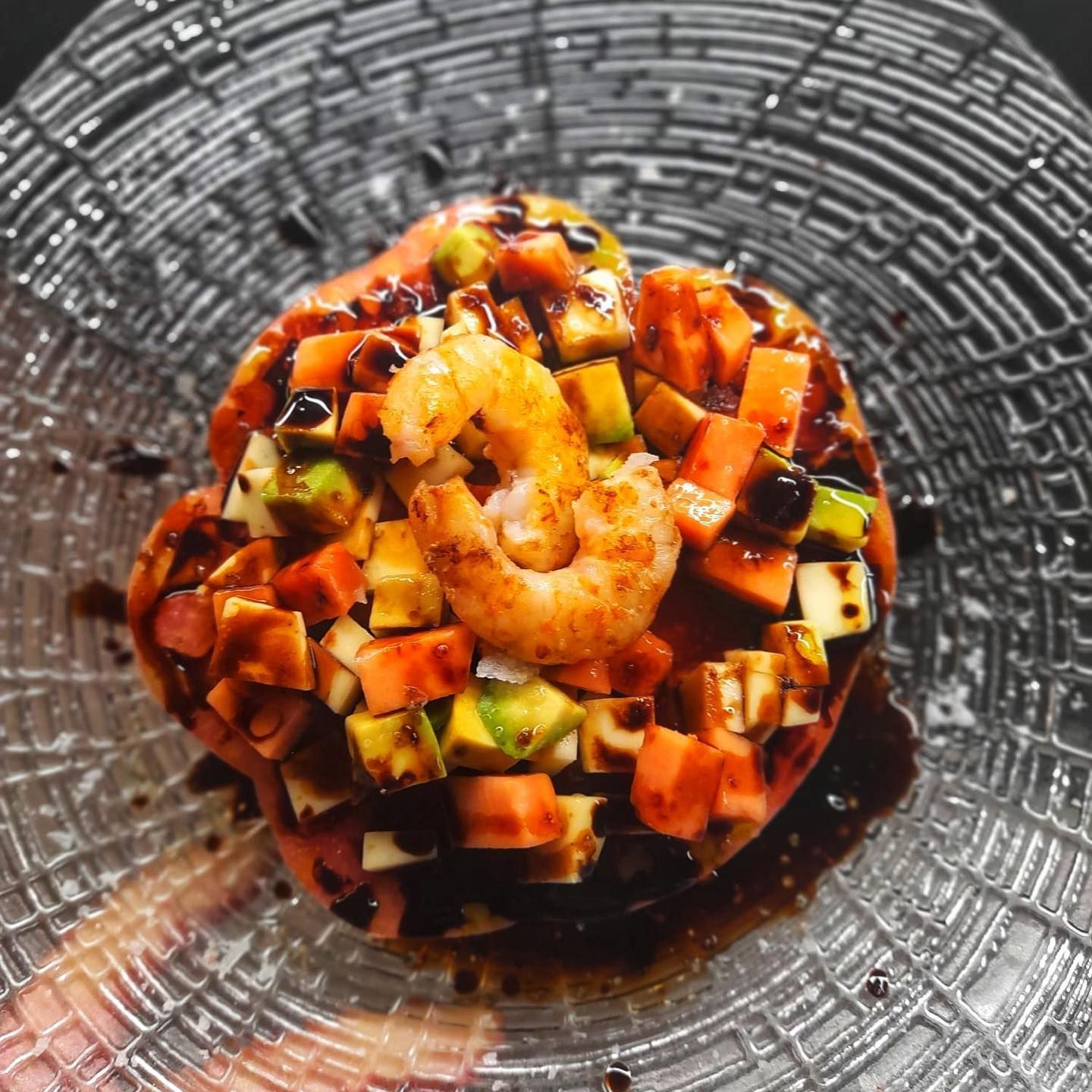 Una gamba cubierta con aguacate en dados y ensalada de tomate, rociada con glaseado balsámico y servida en un plato de cristal texturizado.