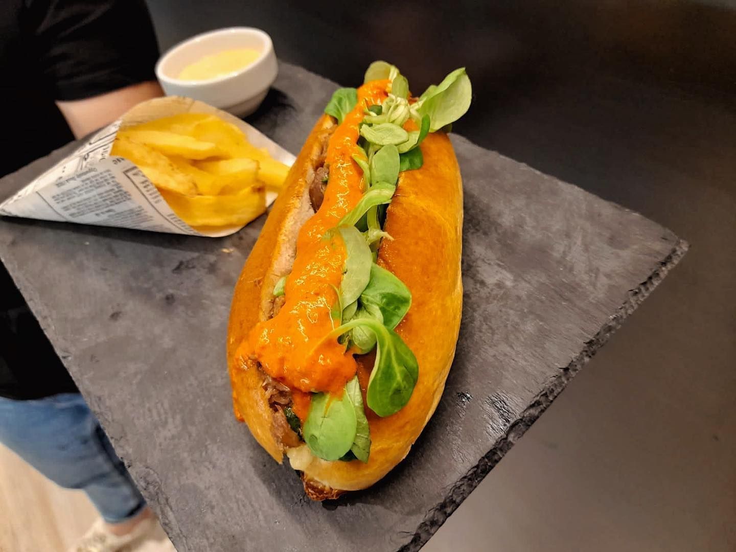 Un sándwich cubierto con verduras y salsa de naranja, servido con patatas fritas en un cono de papel y una pequeña porción de salsa para mojar en una bandeja de pizarra.