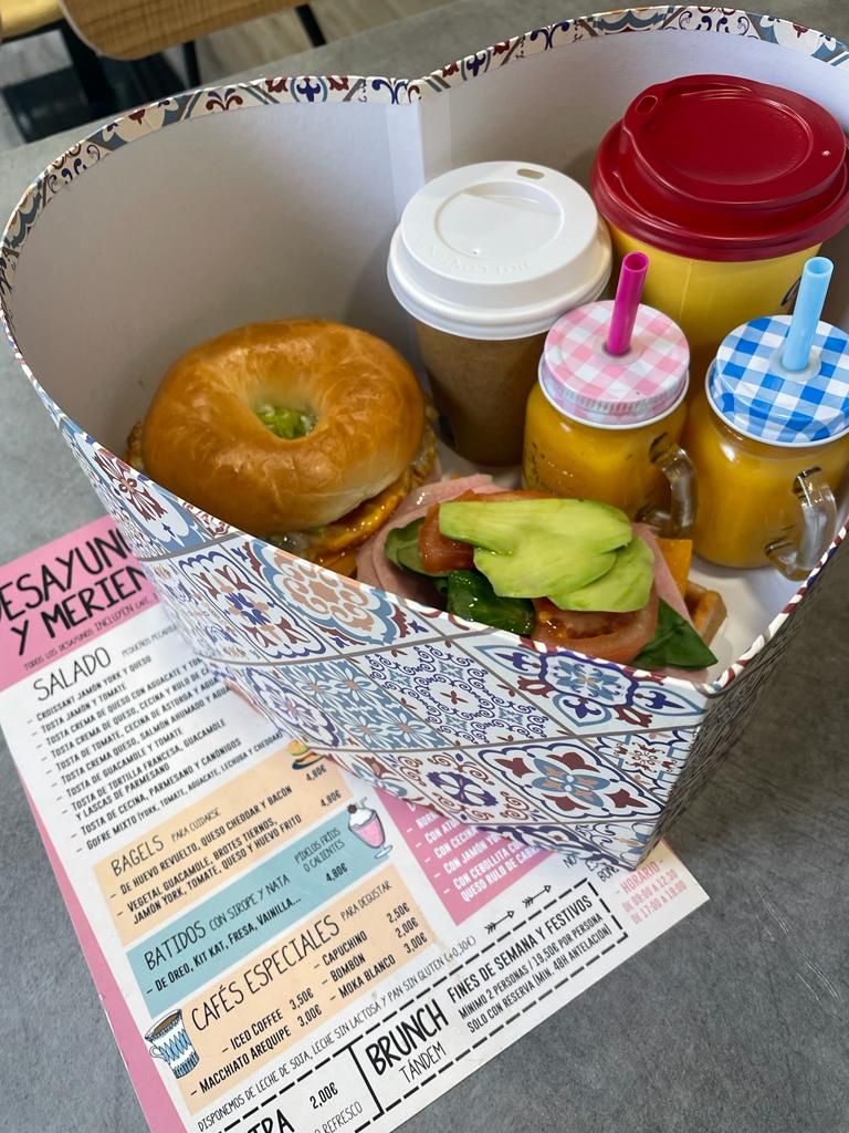 Una caja de regalo con forma de corazón que contiene un bagel, un sándwich de aguacate, dos vasos pequeños de zumo y una taza de café.