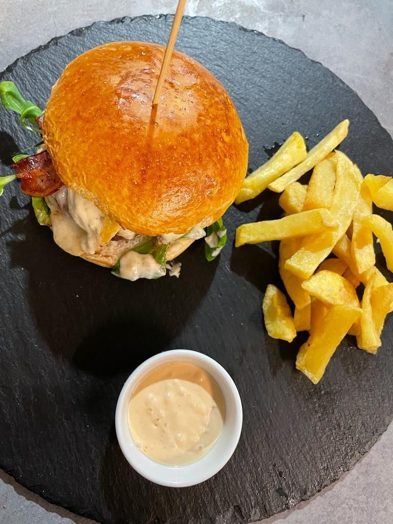 Una hamburguesa con lechuga y salsa, servida con patatas fritas y un pequeño cuenco de salsa para mojar en una pizarra.