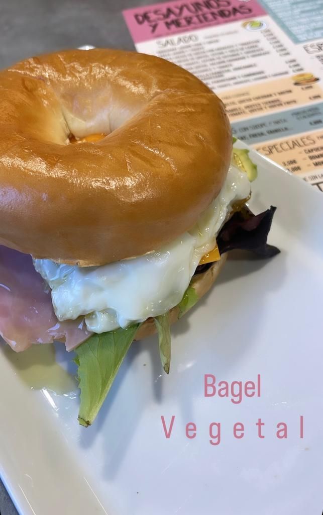 Un sándwich de bagel con jamón, huevo y lechuga en un plato blanco, con un menú borroso al fondo.