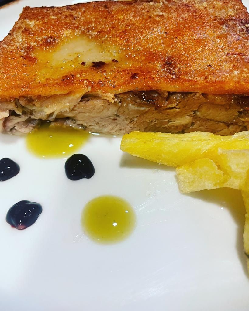 Una loncha de panceta de cerdo con la piel crujiente servida en un plato blanco con bastones de patata frita, aceite de oliva y gotas de salsa de frutos rojos oscuros.