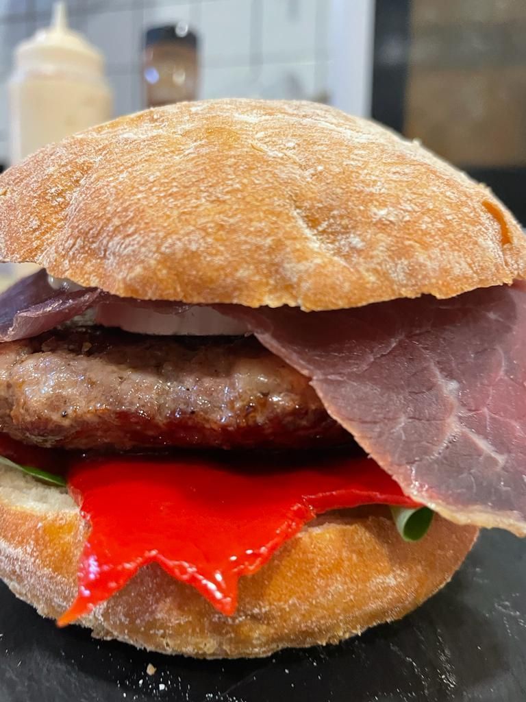 Una hamburguesa en un pan rústico, con una hamburguesa de carne de res, una loncha de embutido y un pimiento rojo brillante sobre una superficie negra.