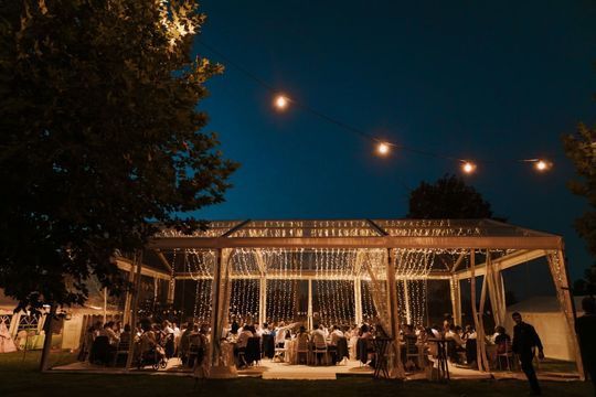 Los invitados se sentaron a una cena bajo un pabellón iluminado al aire libre por la noche, rodeados de árboles y guirnaldas de luces.