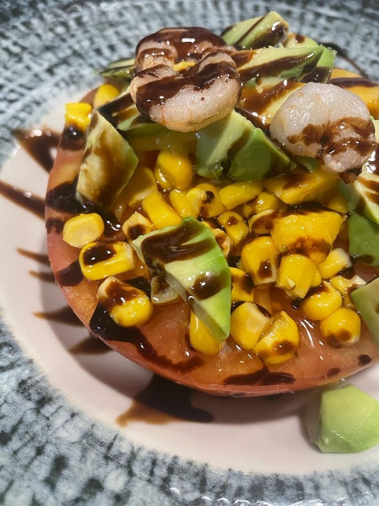 Una rodaja de tomate cubierta con maíz, trozos de aguacate y camarones, rociada con glaseado balsámico en un plato estampado.