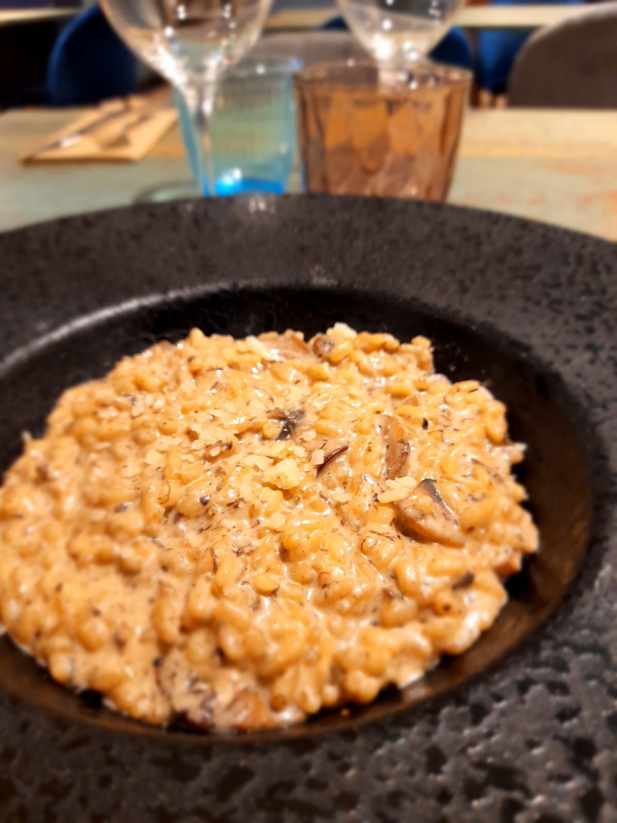 Un bol de risotto cremoso de champiñones servido en un plato negro texturizado, con vasos sobre una mesa de restaurante al fondo.