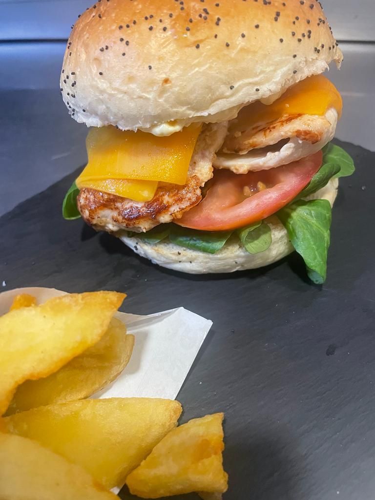 Una hamburguesa de pollo con queso cheddar, tomate y lechuga en un pan con semillas de amapola, servida con papas fritas en un plato oscuro.