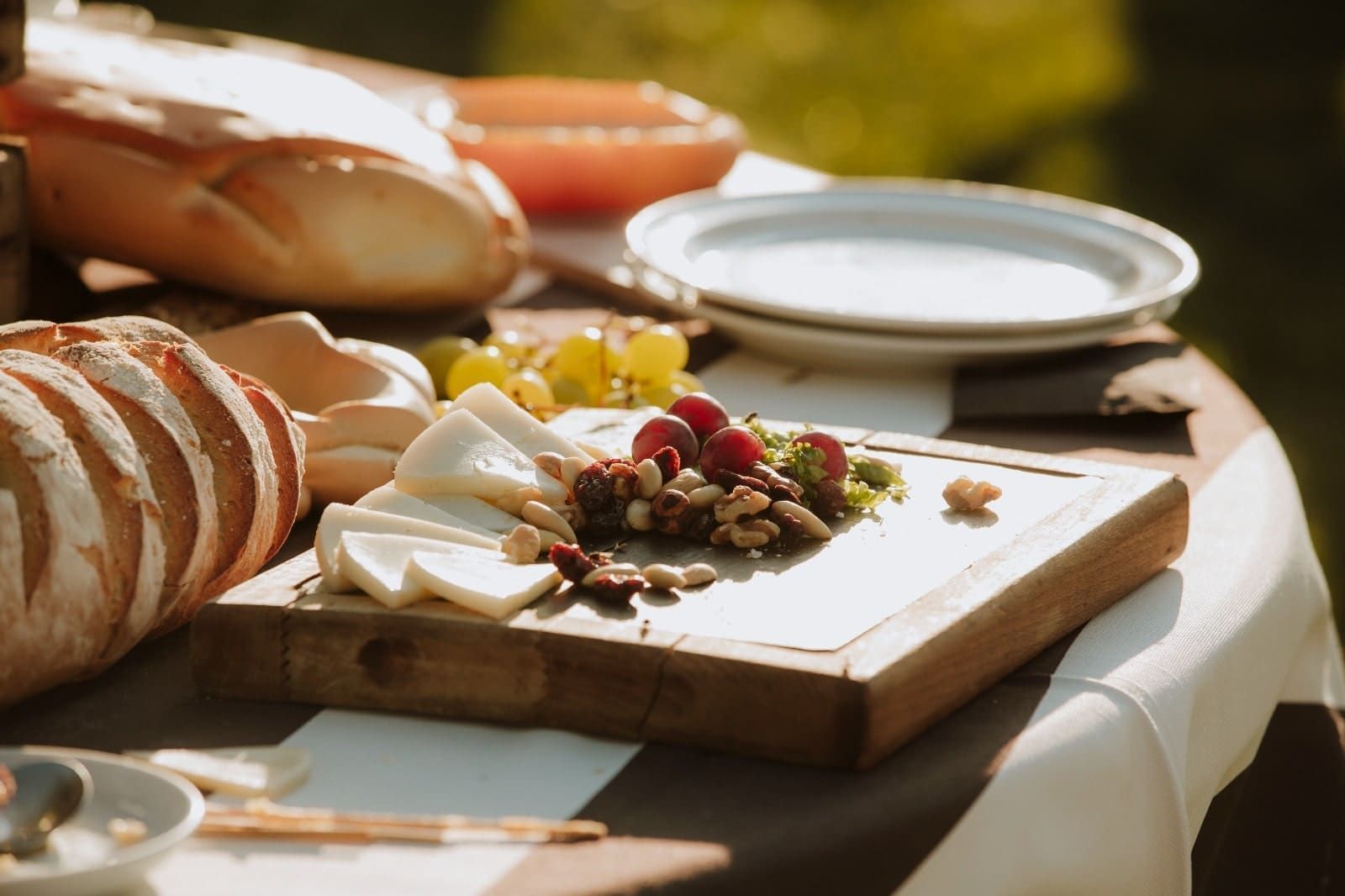 Una tabla de embutidos con queso en lonchas, frutos secos y bayas reposa sobre una mesa junto a barras de pan y platos apilados.