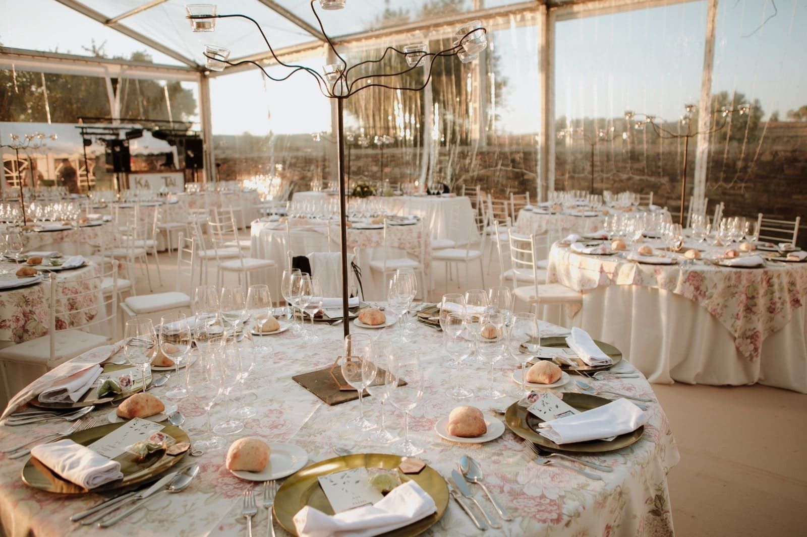 Mesas con manteles florales preparadas para una boda al aire libre dentro de una carpa transparente al atardecer.