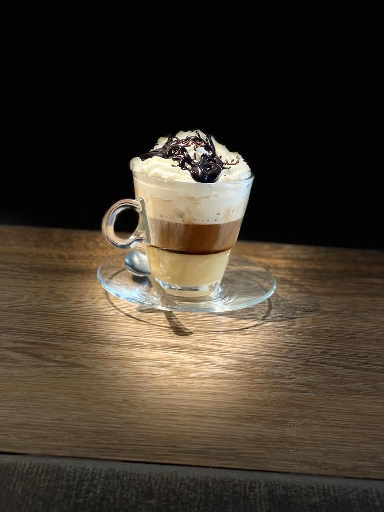 Una taza de cristal con café en capas, coronada con crema batida y sirope de chocolate, que reposa sobre un platillo en una superficie de madera.