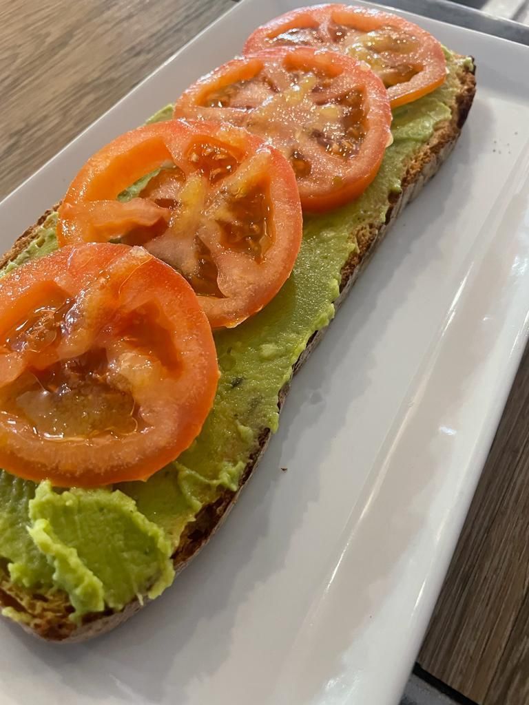 Una rebanada de pan de masa madre tostado, cubierta con aguacate triturado y cuatro rodajas finas de tomate rojo fresco, servida en un plato blanco.