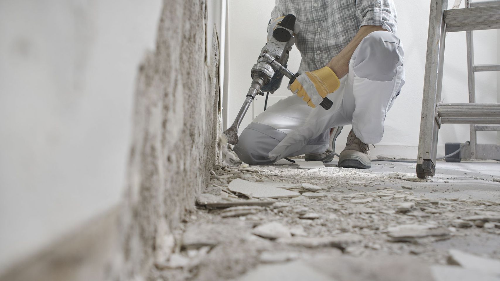 Un ouvrier du bâtiment perce un mur lors de travaux de rénovation ; de la poussière et des débris jonchent le sol.