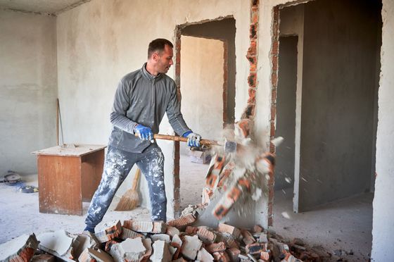 Un ouvrier démolit un mur à coups de masse au milieu des décombres d'une pièce inachevée.