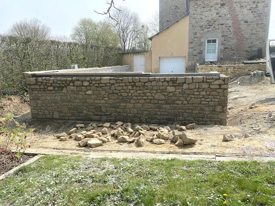 Mur de soutènement en pierre à côté d'une maison, avec des pierres empilées et une pelouse clairsemée dans la cour.