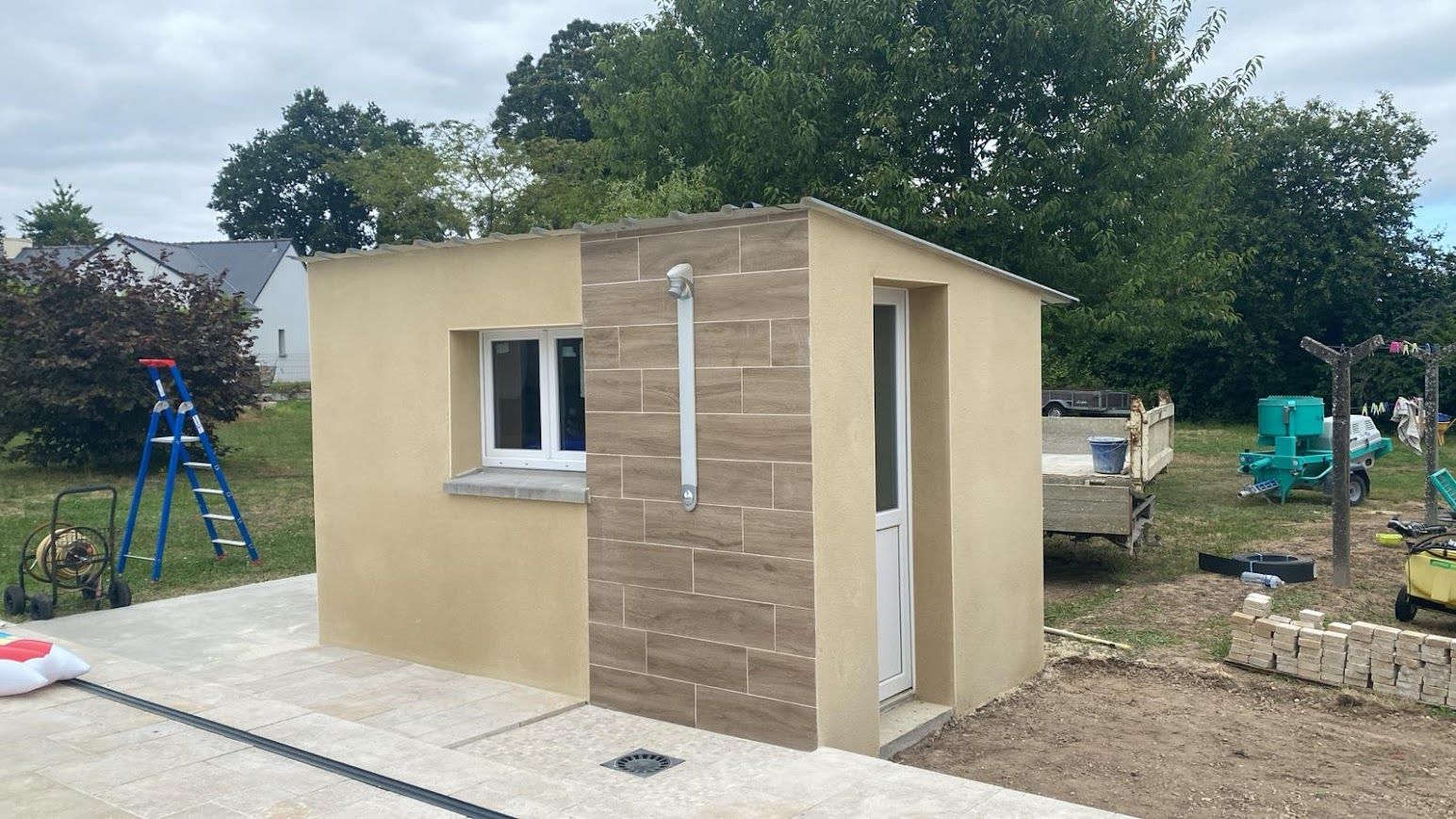 Petit abri en construction sur une dalle de béton dans une cour, avec des arbres et du matériel à proximité.