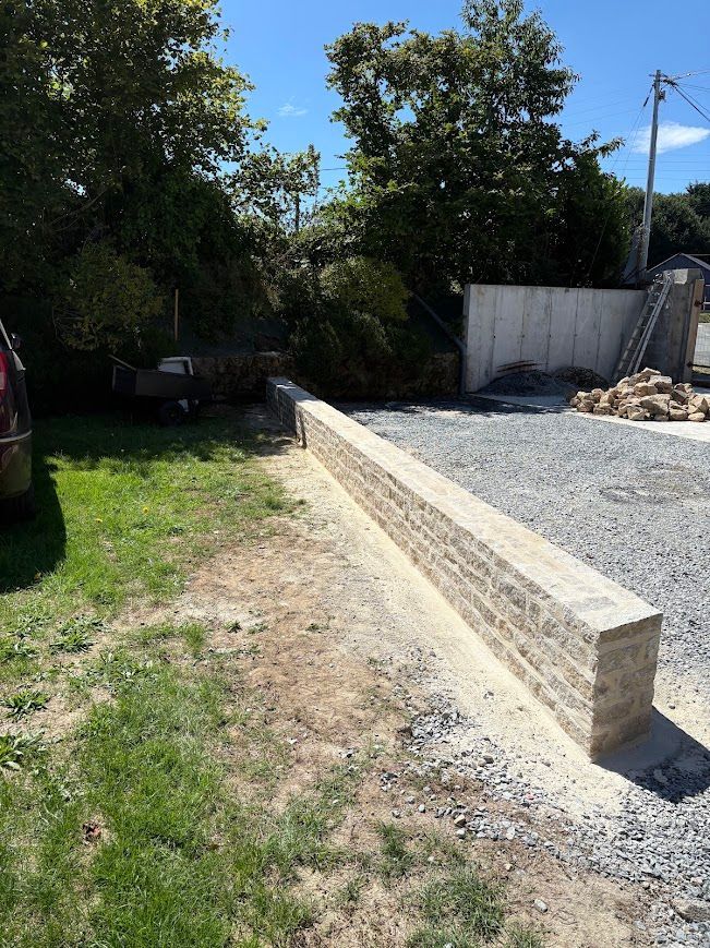 Allée extérieure en gravier longeant un mur de soutènement en bois et une pelouse, avec des arbres et un mur en béton en arrière-plan.