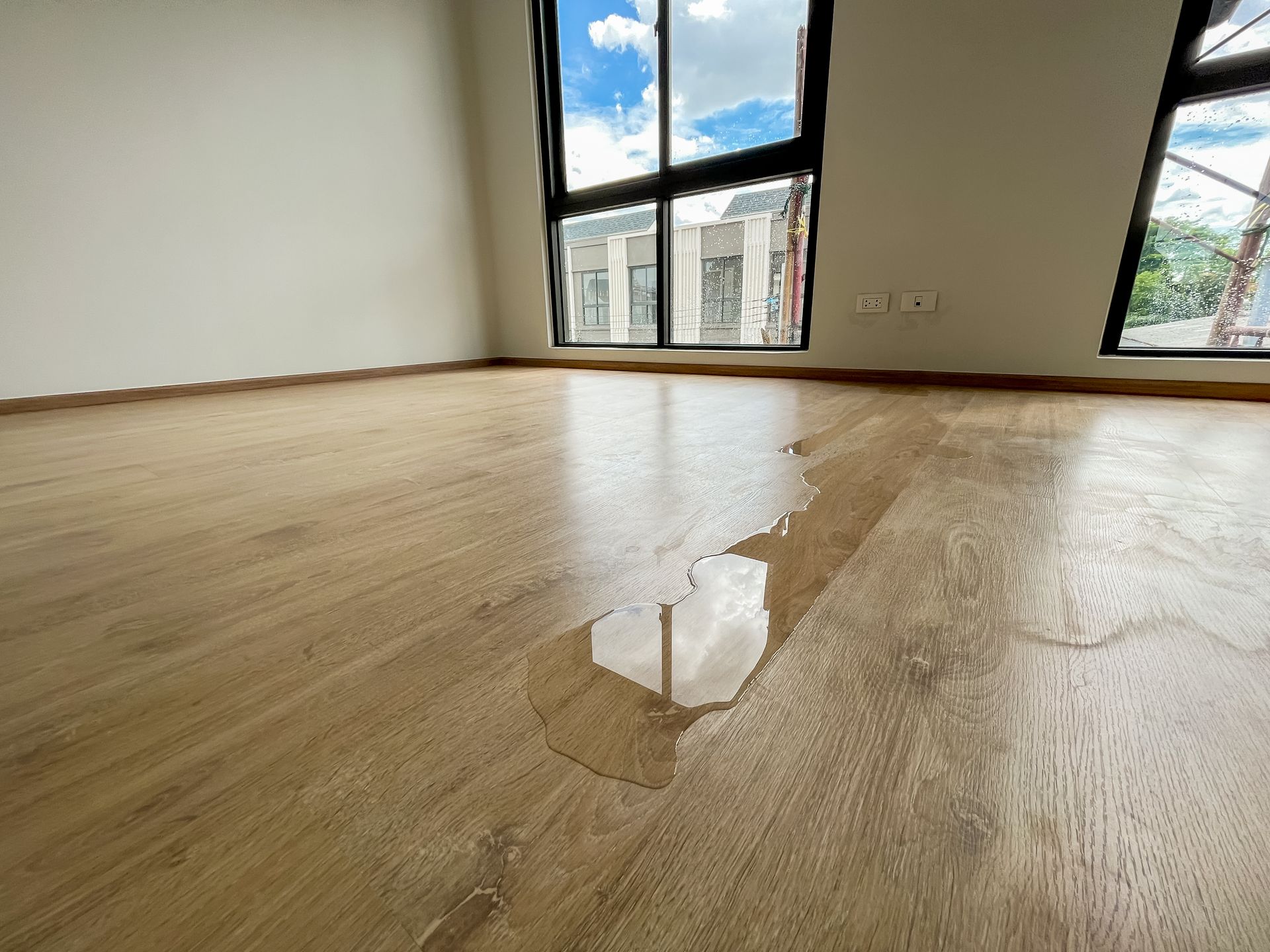 De l'eau sur le parquet d'une pièce vide, près de fenêtres offrant une vue partielle sur les bâtiments extérieurs.