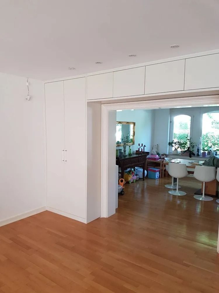 Leerer, weißer Raum mit Holzboden und einem großen offenen Durchgang zu einem hellen Essbereich
