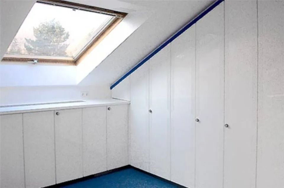 Weißer Dachbodenraum mit schräger Decke, Dachfenster, Einbauschränken und blauem Teppichboden