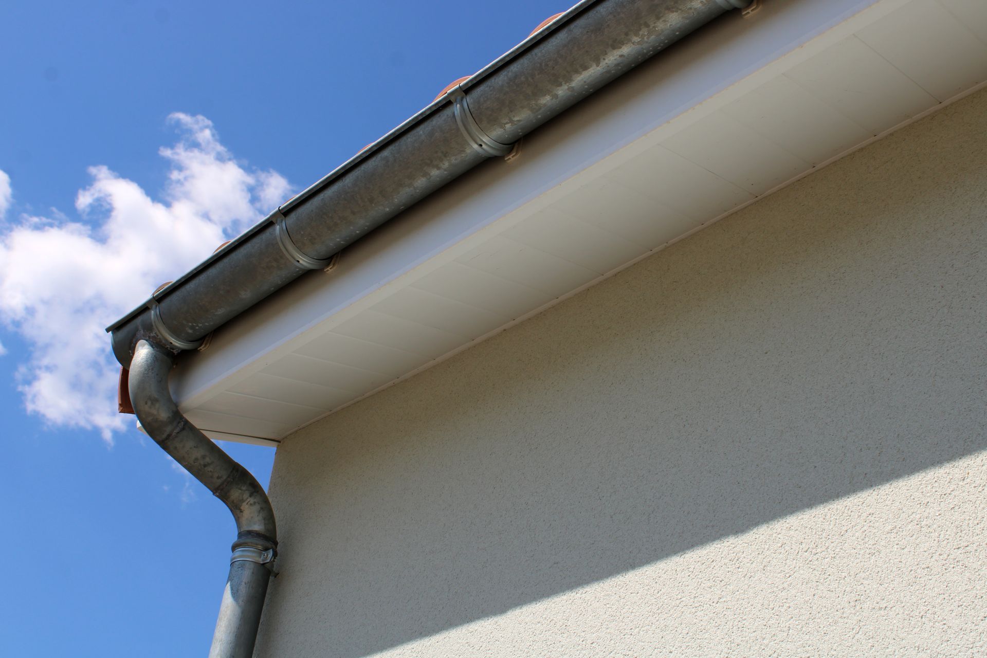 Gouttière et descente pluviale sur une maison à la façade beige, sur fond de ciel bleu nuageux.