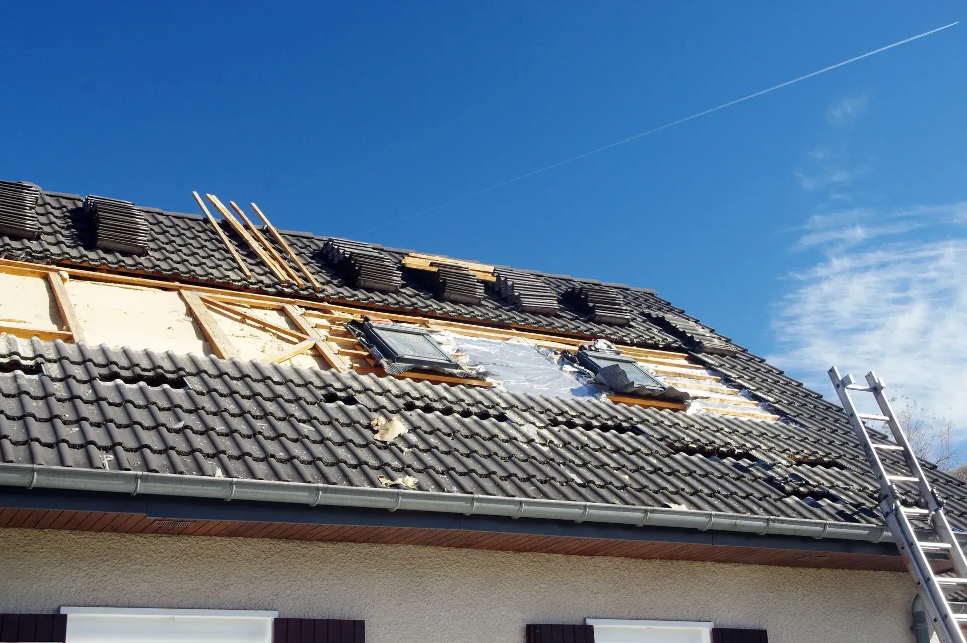 Toiture en cours de réparation, tuiles partiellement retirées, laissant apparaître la charpente en bois, une échelle et des puits de lumière sous un ciel bleu.