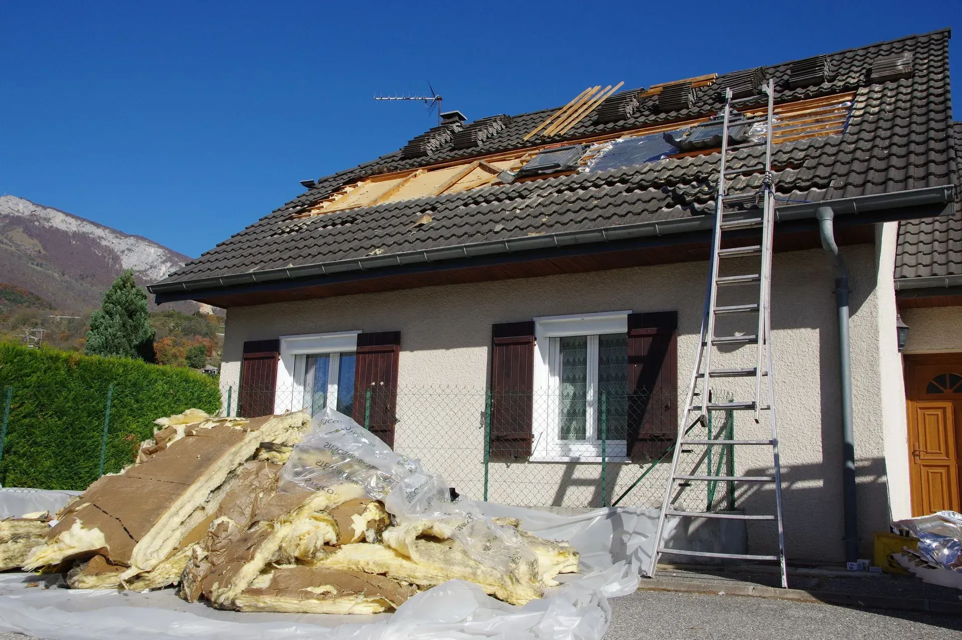 Maison avec toit endommagé et isolation au sol, échelle appuyée contre le mur.