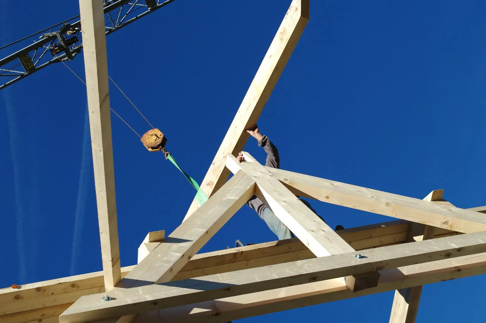 Un ouvrier du bâtiment positionne une poutre en bois sur une charpente de toit, une grue est visible en arrière-plan, sous un ciel bleu éclatant.