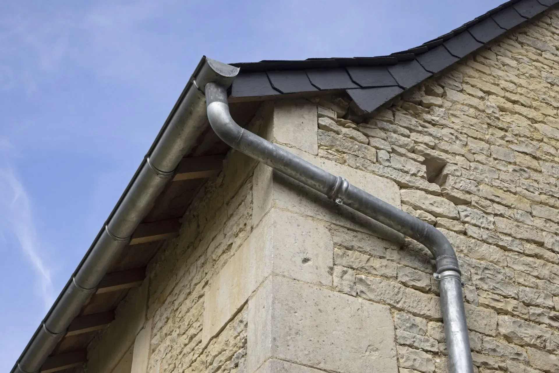 Gouttière métallique fixée à un bâtiment en pierre avec un toit en bardeaux foncés.