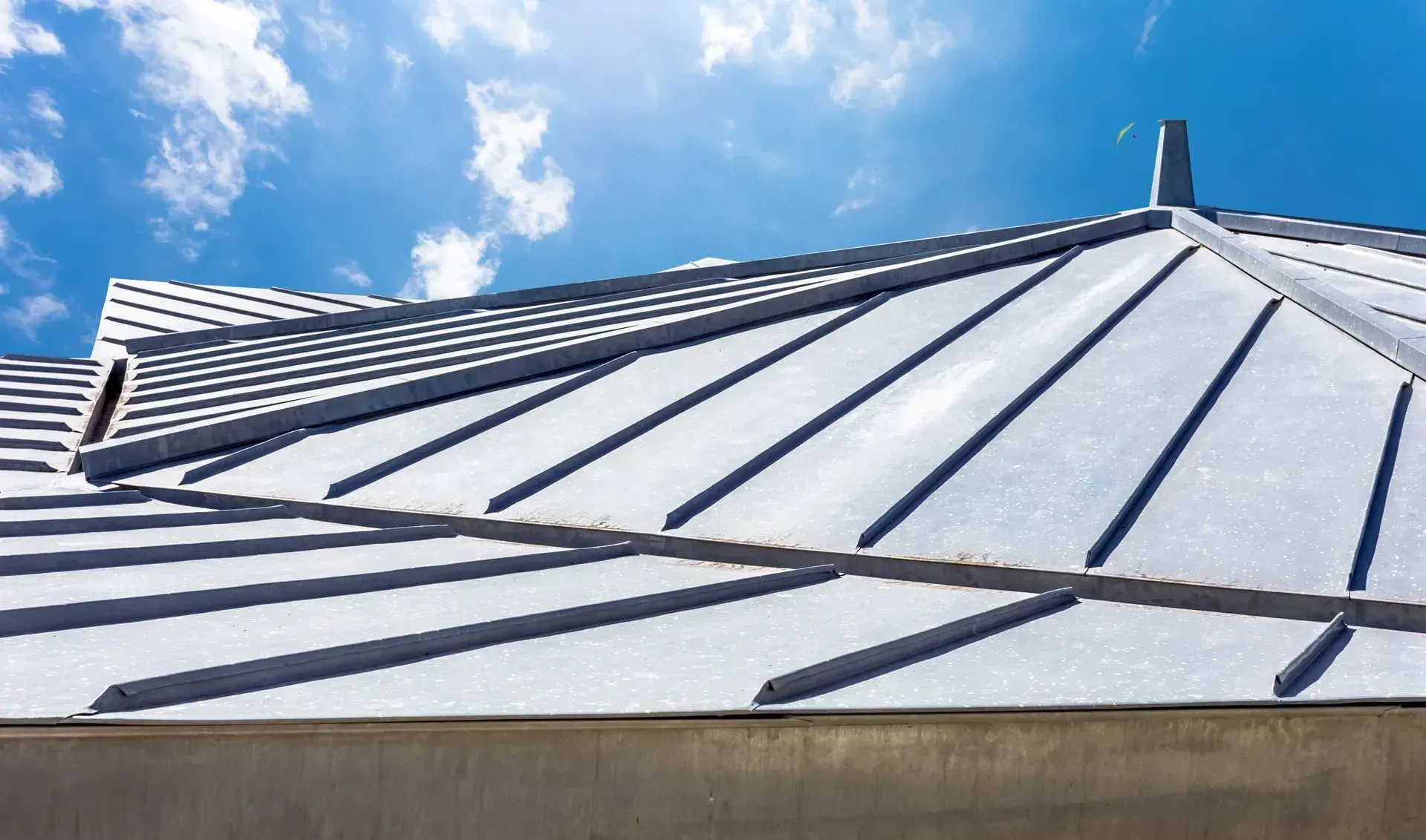 Gros plan sur une toiture moderne en métal gris à joints debout se détachant sur un ciel bleu vif parsemé de nuages ​​épars.