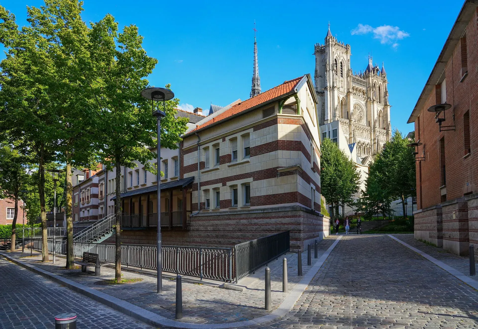 Rue pavée avec des bâtiments et une cathédrale en arrière-plan, sous un ciel bleu.