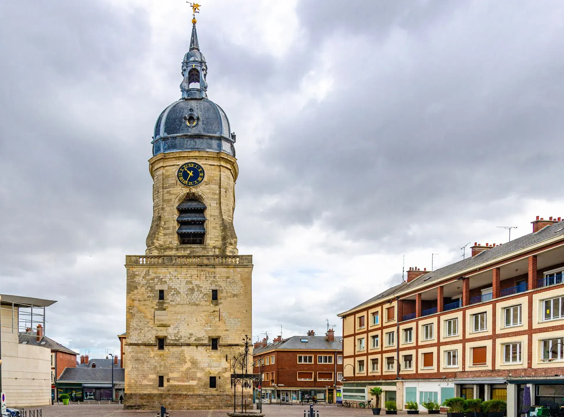 Tour de l'horloge sur une place de ville, avec des bâtiments sous un ciel nuageux.