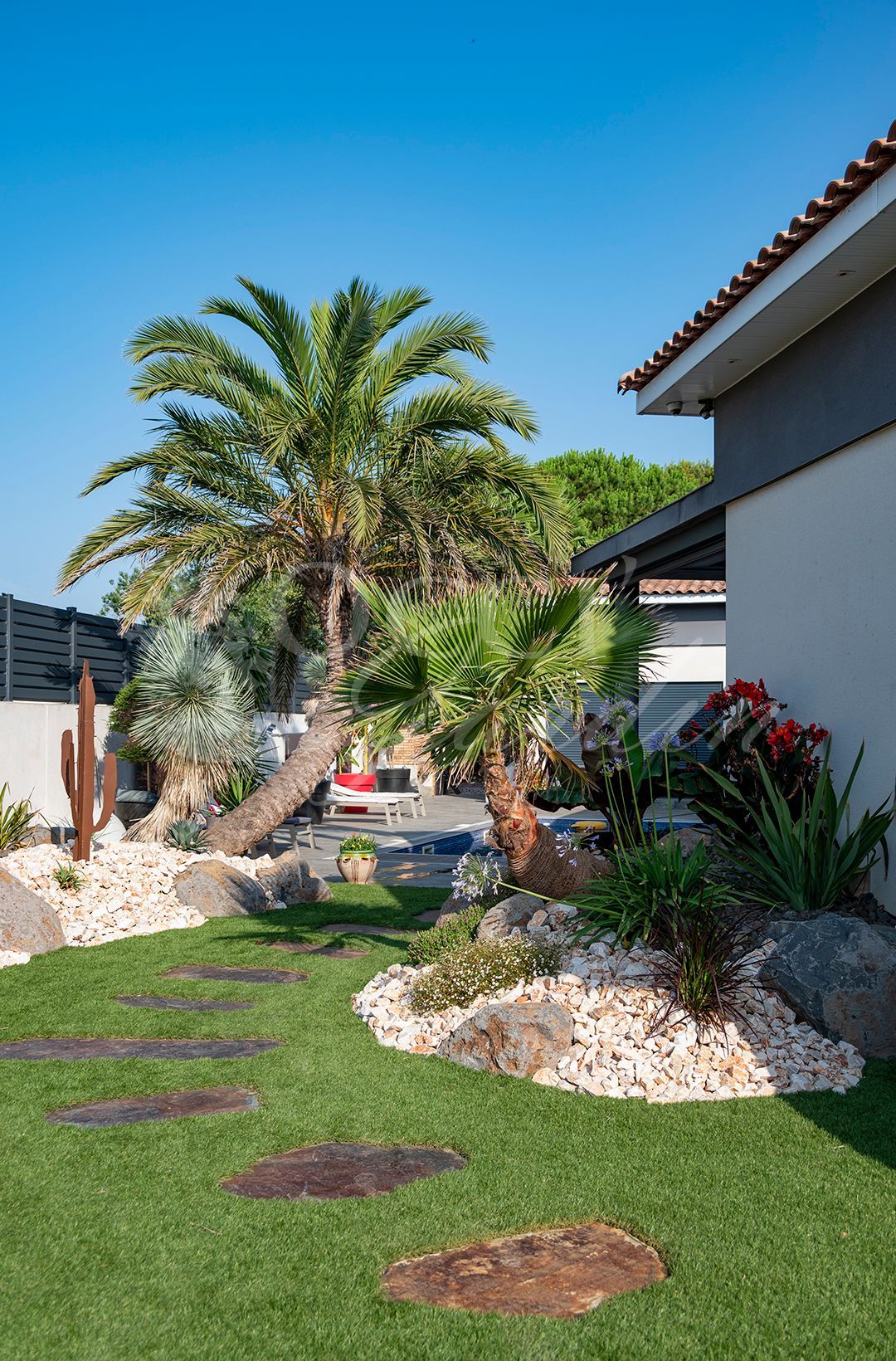 Un jardin luxuriant avec des palmiers, un jardin de rocaille et des pas japonais menant à une maison sous un ciel bleu azur.