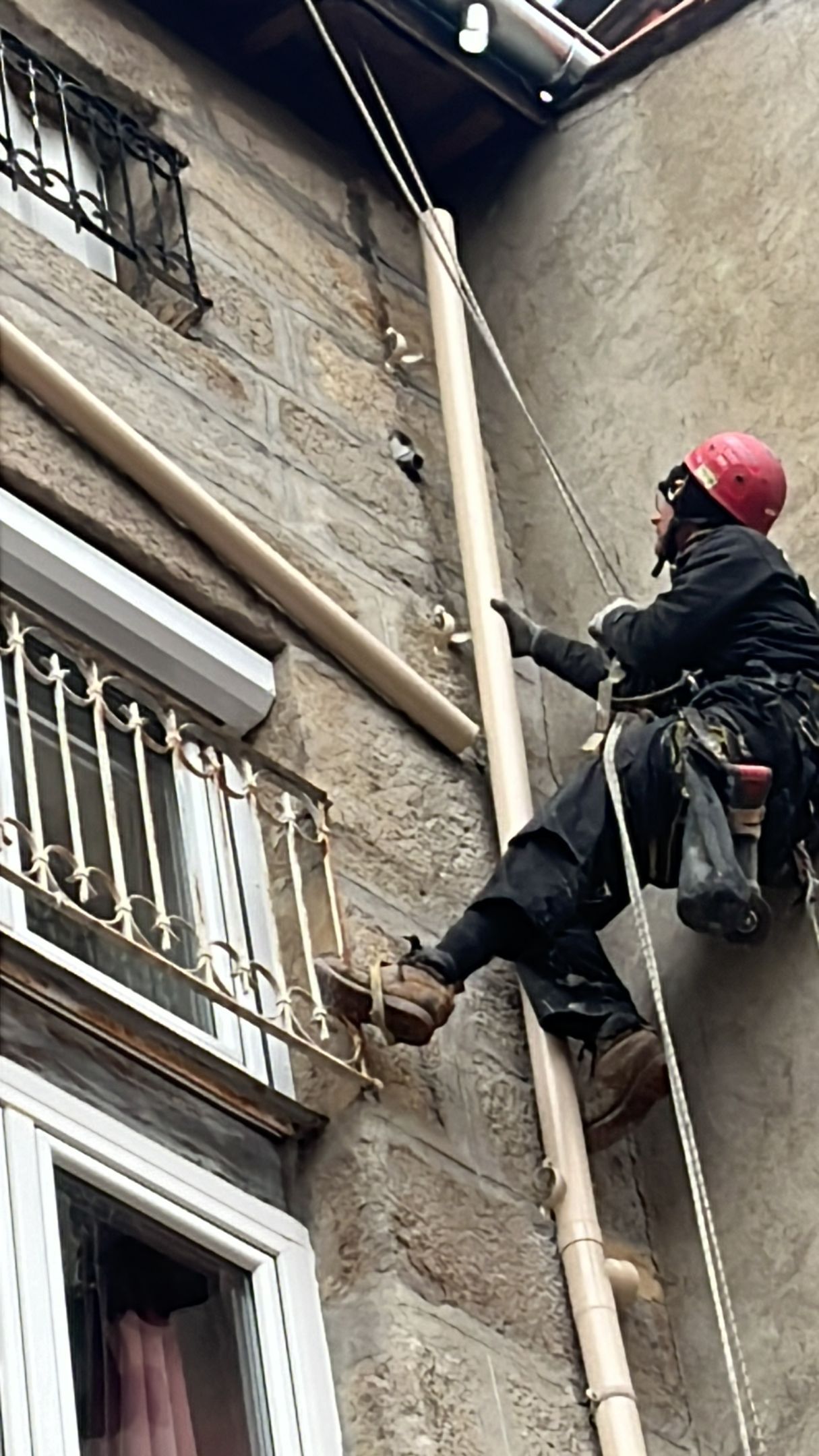 Un ouvrier portant un casque rouge grimpe à une corde sur la façade d'un bâtiment en brique.