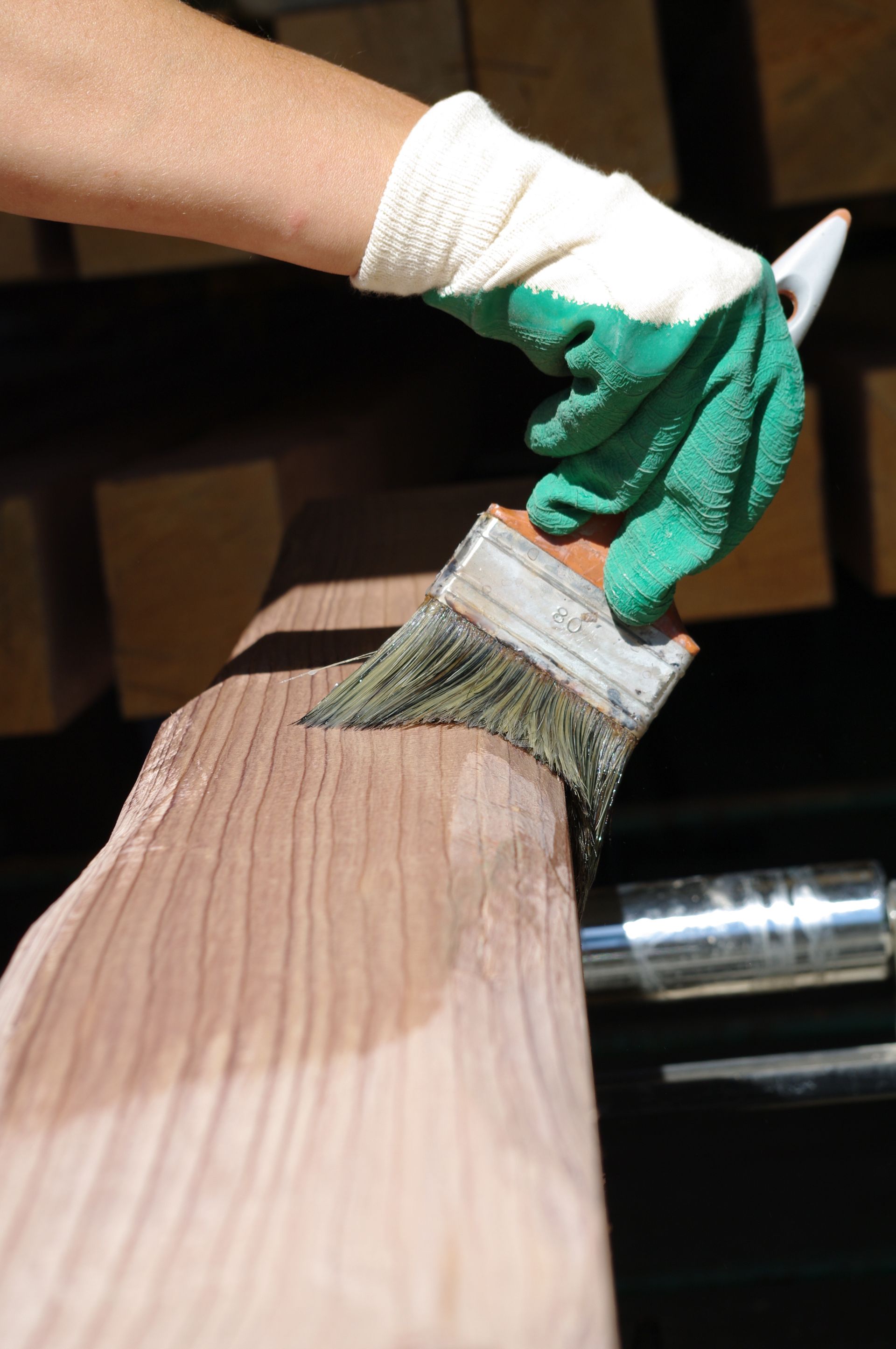 Gros plan sur une main avec un gant vert tenant un pinceau et appliquant une finition transparente sur une surface en bois.