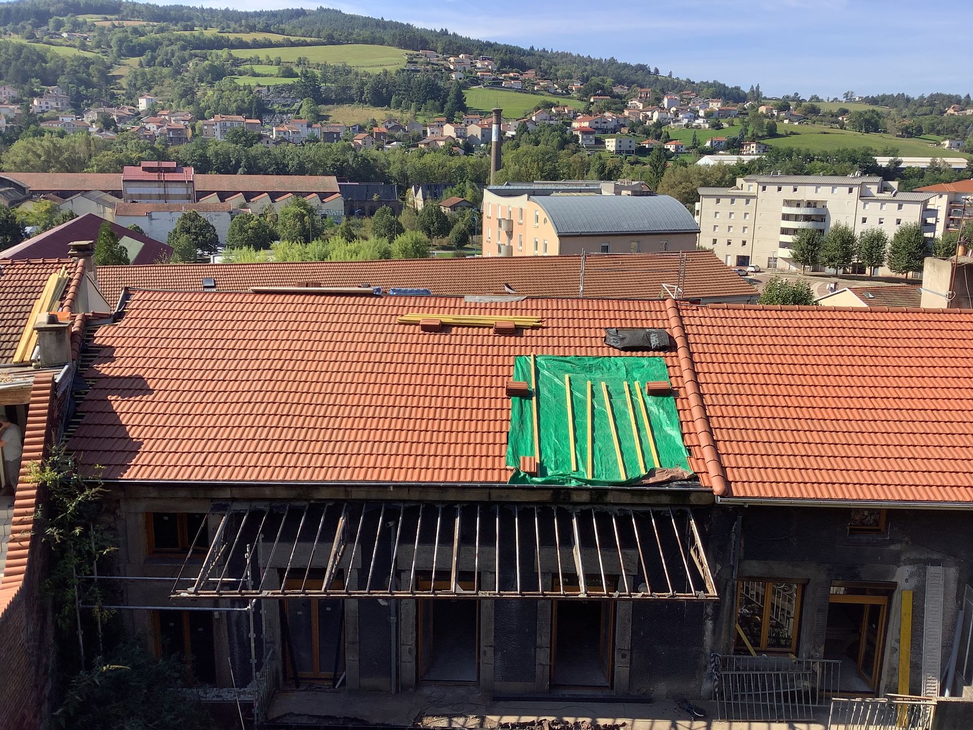 Toiture en réparation, recouverte de tuiles rouges et d'une bâche verte, donnant sur une ville à flanc de colline et des champs.