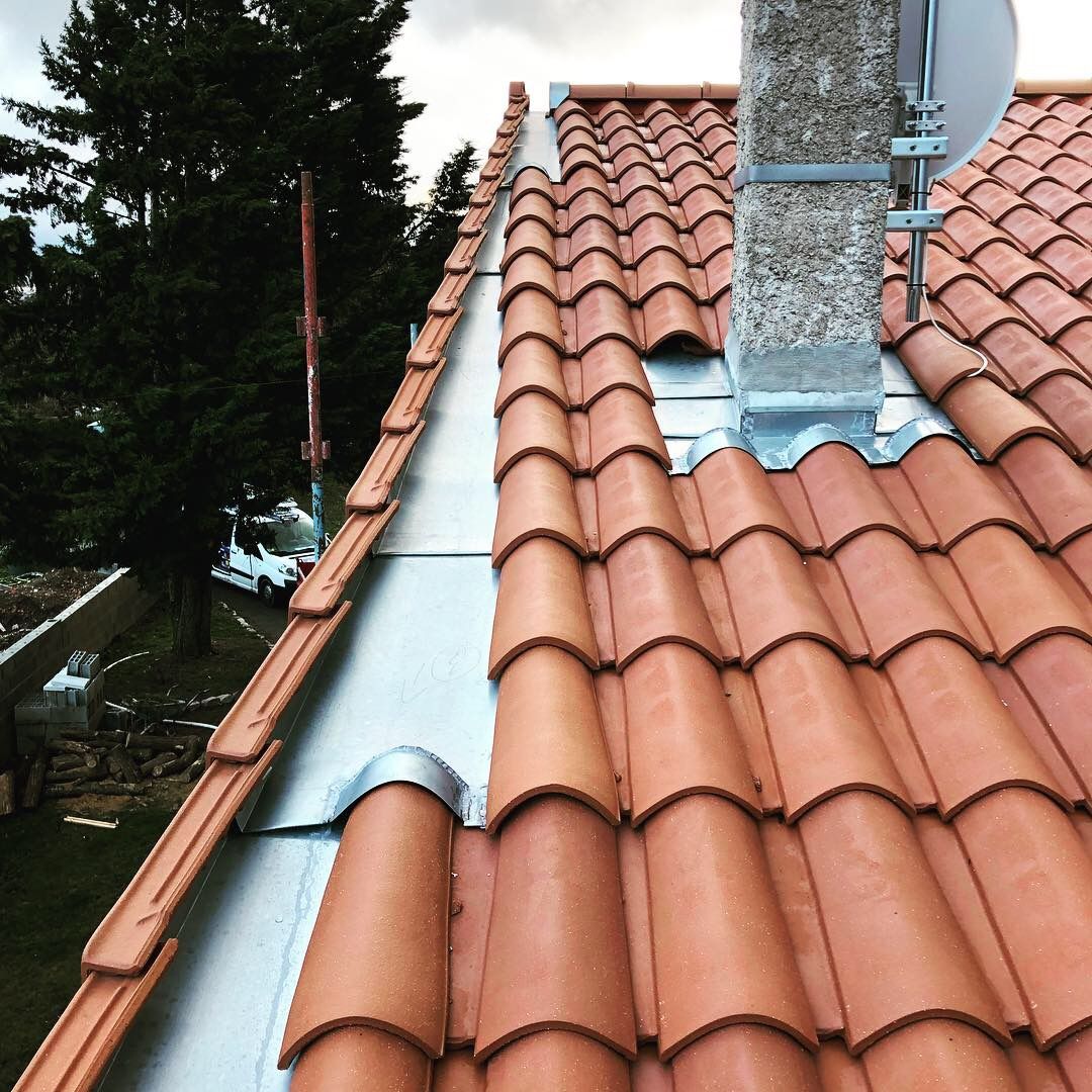 Gros plan sur un toit de tuile en terre cuite rouge avec une bande de solin métallique et une cheminée en pierre.