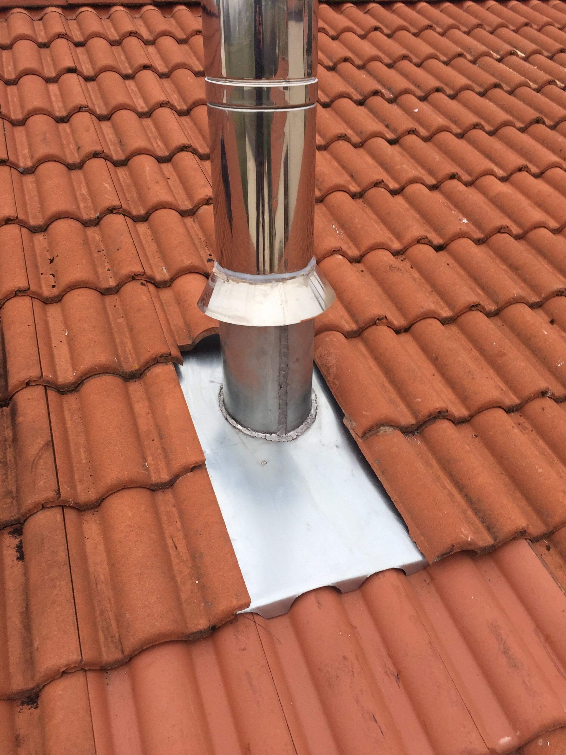 Conduit de cheminée métallique sur un toit de tuilesrouge, avec un collier de solin blanc autour.