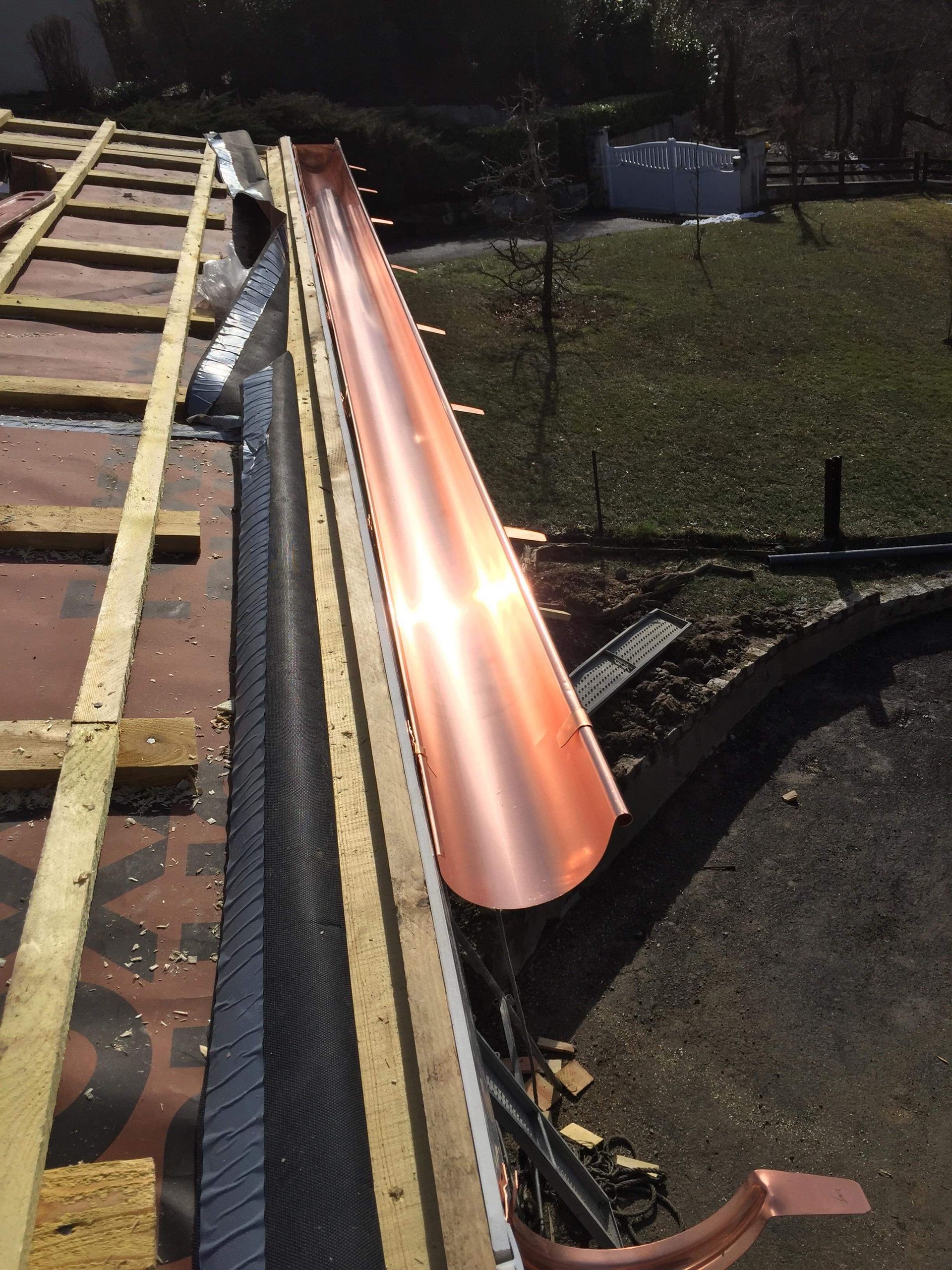 Une nouvelle gouttière en cuivre installée en bordure d'un toit en construction, laissant apparaître les liteaux en bois et la sous-couche de toiture.