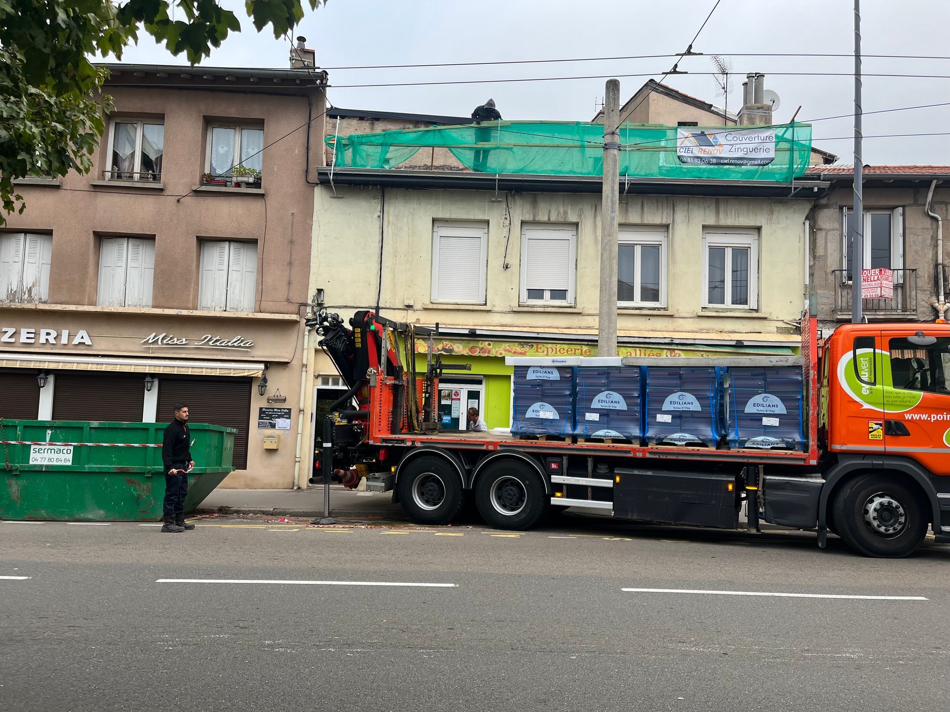 Un camion orange transportant des caisses bleues est stationné dans une rue, à côté de vieilles devantures de magasins.