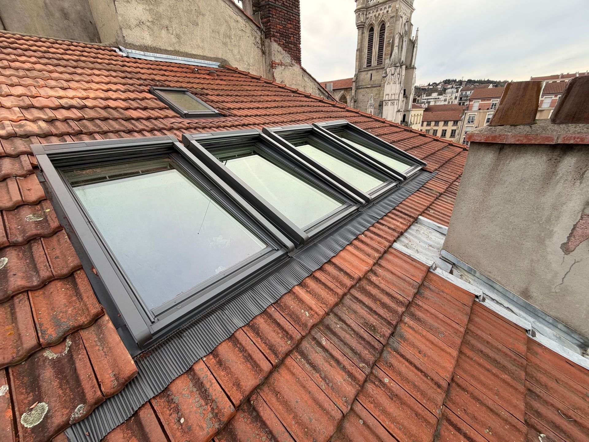 Trois puits de lumière installés dans un toit de tuiles, à côté d'une grande cheminée, donnant sur une ville européenne avec un clocher.