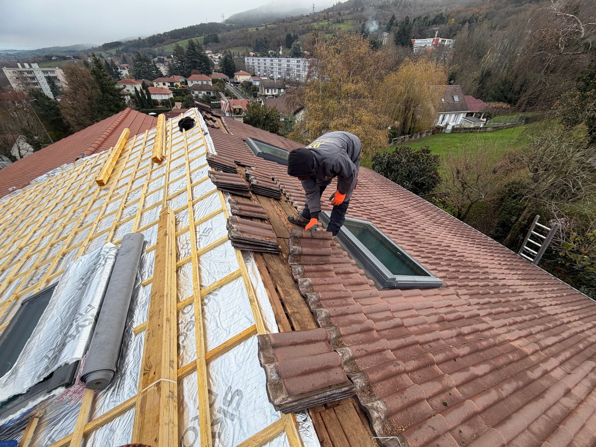 Ouvrier travaillant sur un toit en tuile, installant de l'isolant à côté d'un puits de lumière.