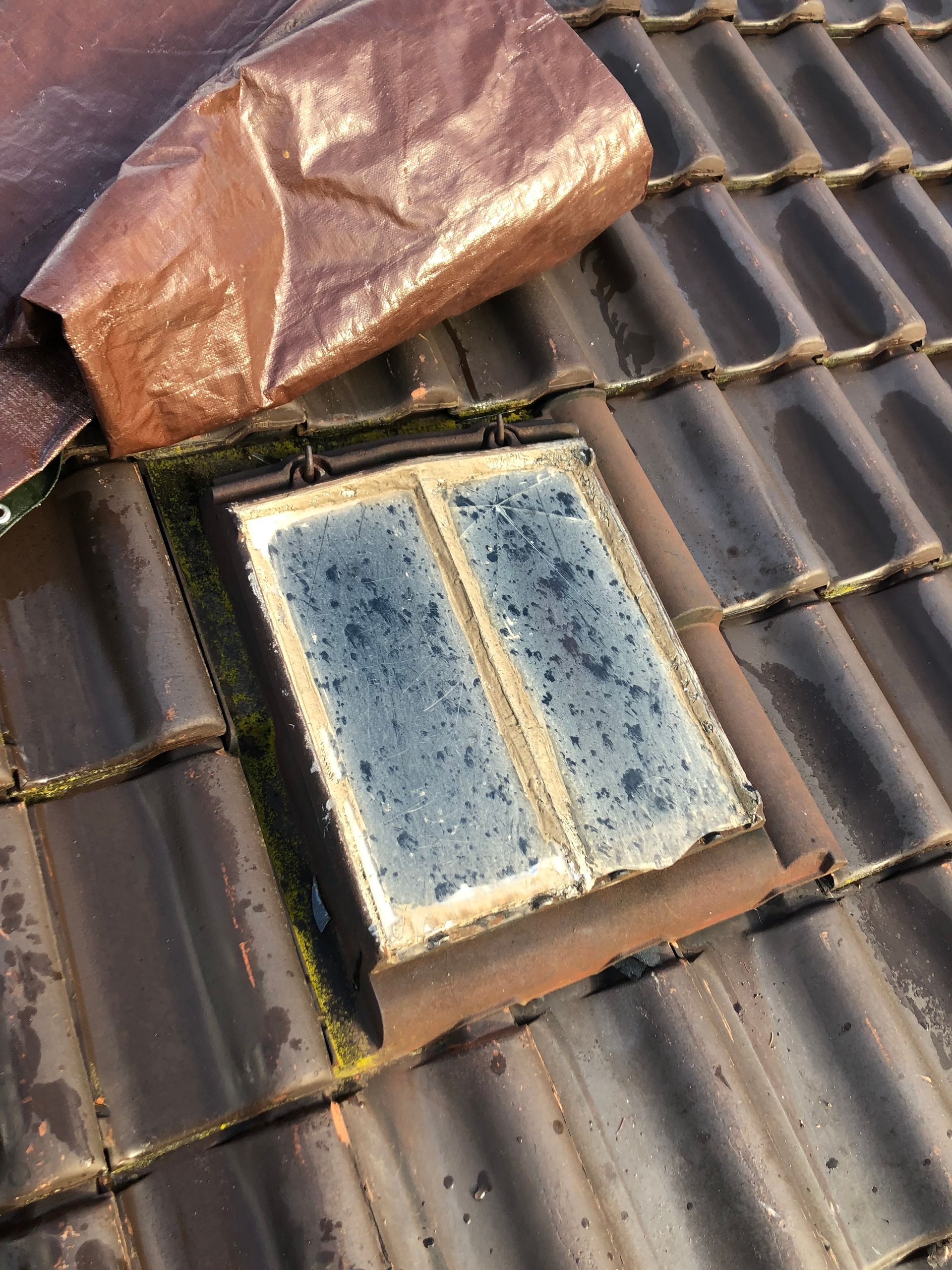 Puits de lumière ouvert sur un toit de tuiles brunes avec une tôle de cuivre non fixée au-dessus.