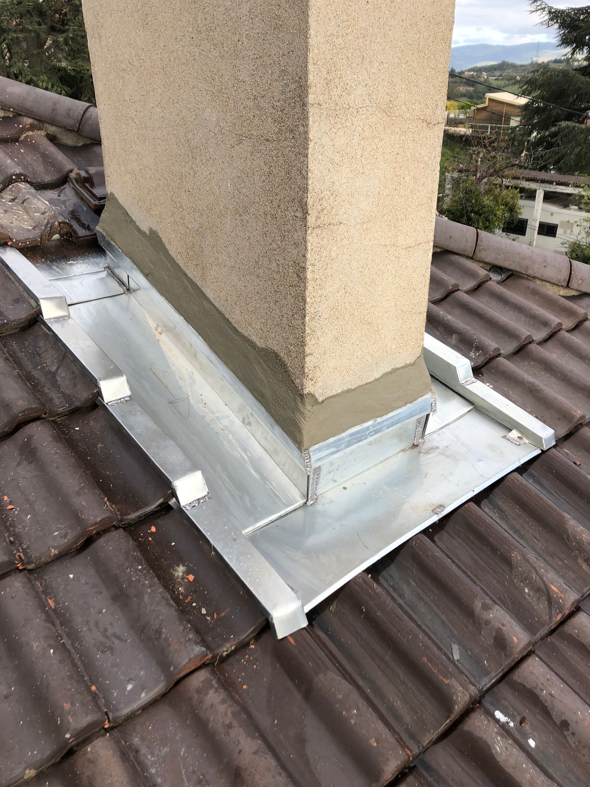 Solins métalliques autour d'une base de cheminée en stuc sur un toit de tuiles, avec des réparations d'étanchéité de toiture visibles.