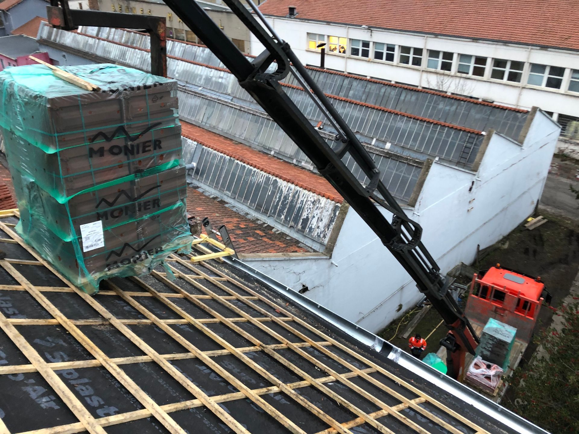 Une grue soulève une palette emballée et la dépose sur le toit d'un chantier de construction où des ouvriers se trouvent en dessous.