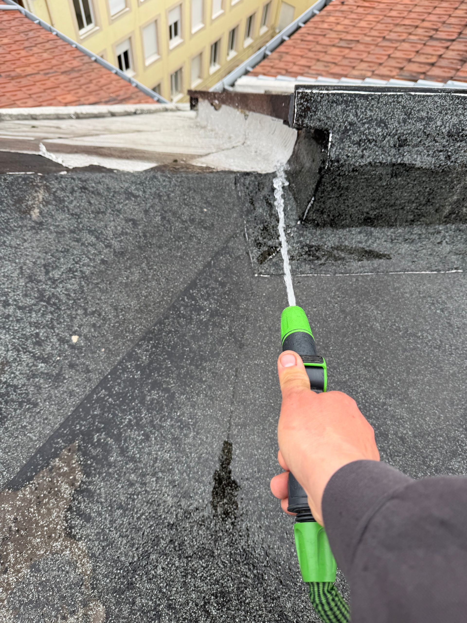 Grattage manuel des joints de toiture à l'aide d'un outil à manche vert, près d'un mur et de bardeaux.