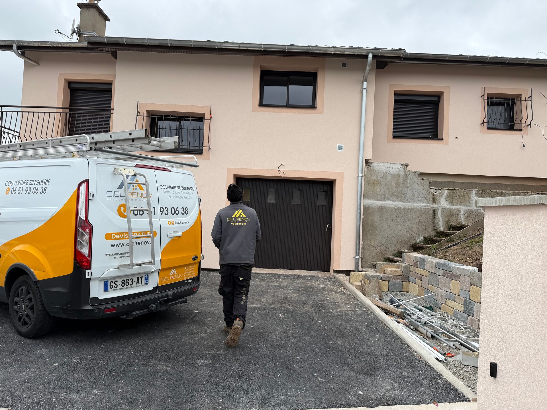 Un ouvrier se dirige vers un garage à côté d'une camionnette utilitaire blanche devant une maison beige.